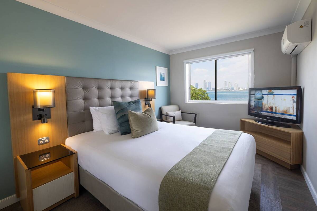 Ein Hotelzimmer mit einem großen Bett, einem Fernseher, einem Fenster mit Blick auf die Stadt und einem Nachttisch mit Lampe.