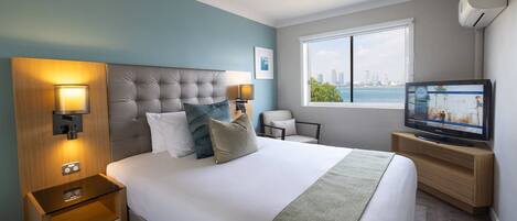 A hotel room with a large bed, a television, a window with a city view, and a nightstand with a lamp.