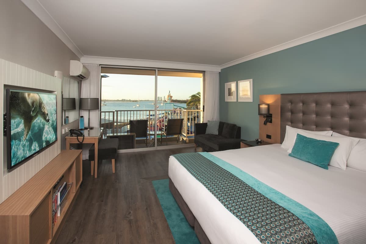 A hotel room with a large bed, a TV, a desk, and a balcony with a view of the sea and buildings.