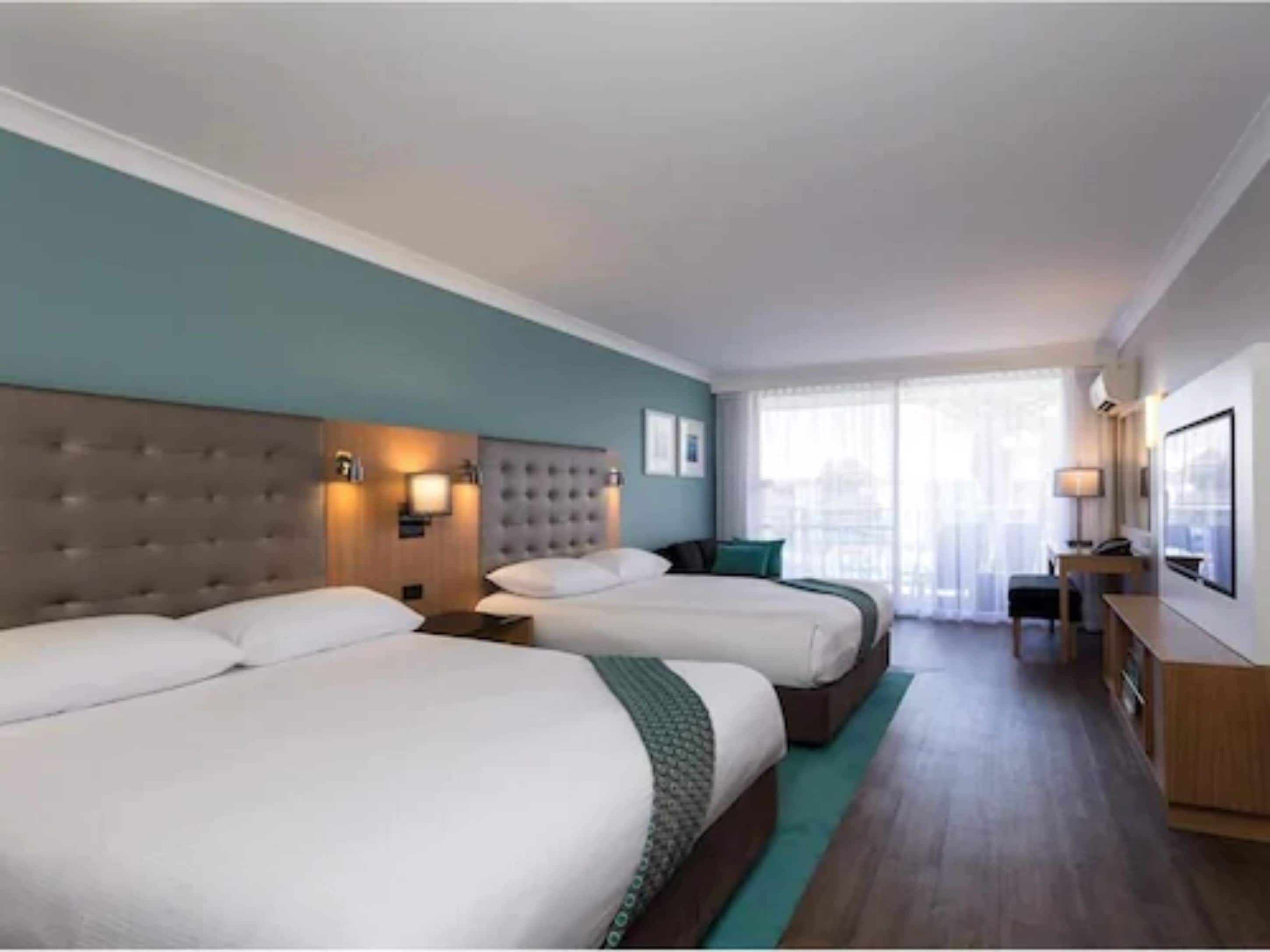 Habitación de hotel con dos camas, un escritorio y vistas de la ciudad a través de amplios ventanales.