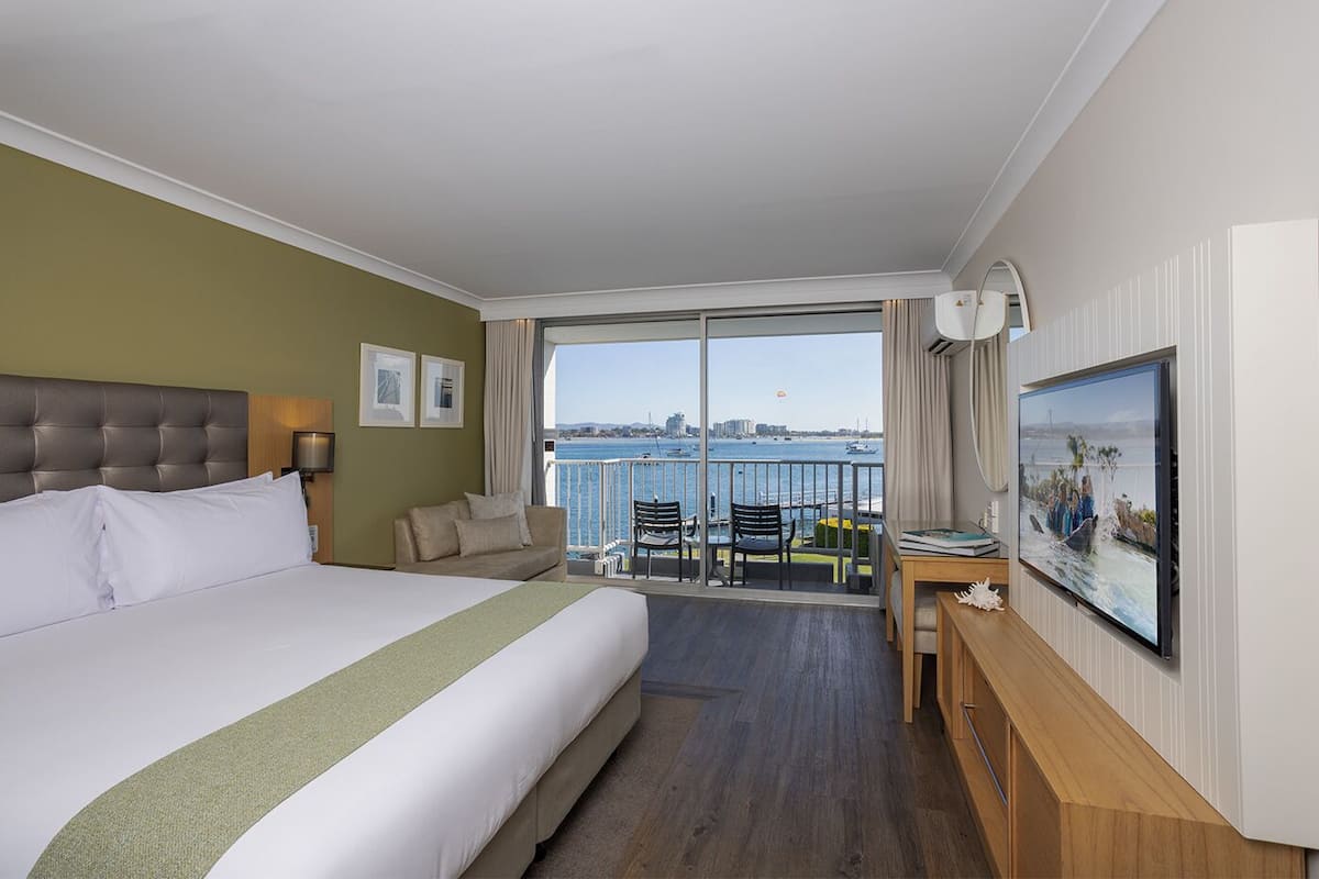 A hotel room with a large bed, a balcony with a view of the water, a desk, and a TV.