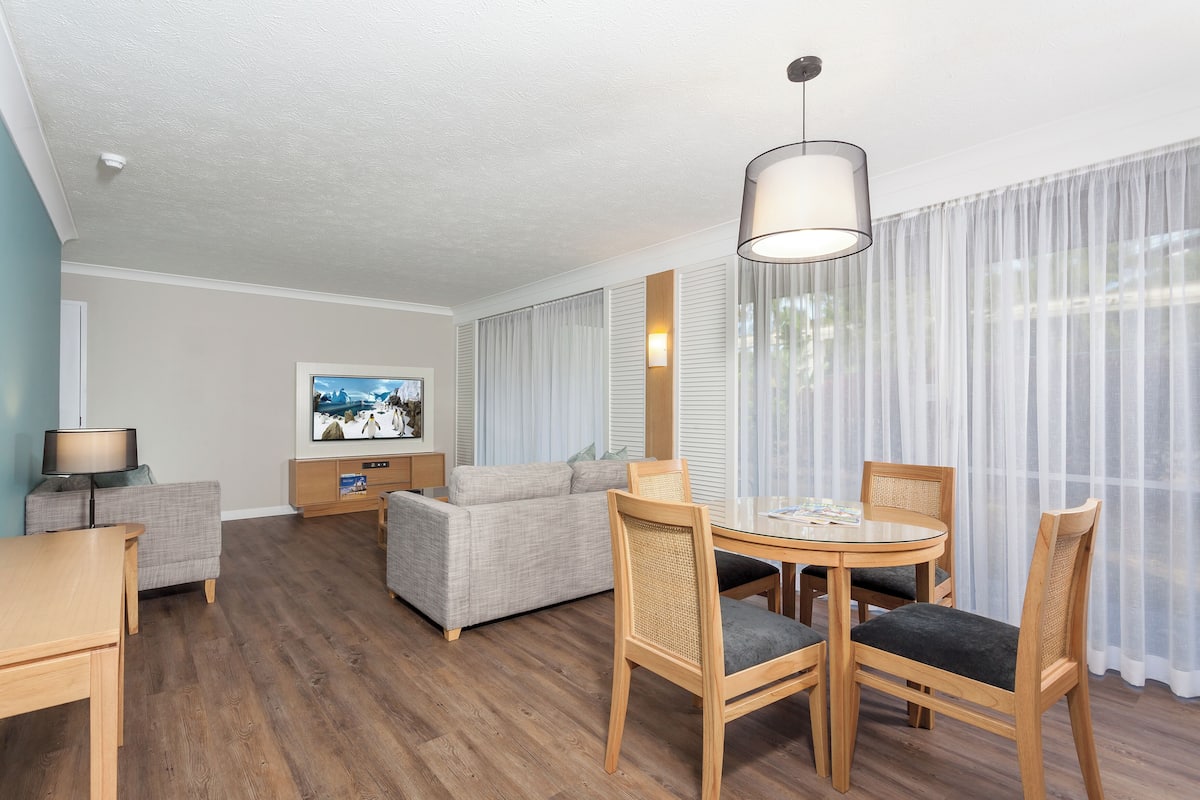 Ein Hotelzimmer mit einem Holztisch, zwei Stühlen, einem Sofa, einem Fernseher und einem großen Fenster mit durchsichtigen Vorhängen.