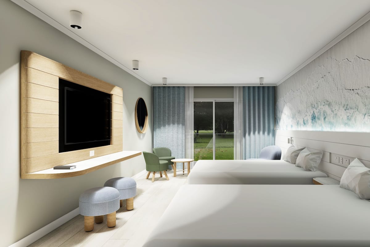 A modern hotel room with a large bed, a TV mounted on the wall, a small seating area with a table and chairs, and a view of a grassy area through the window.