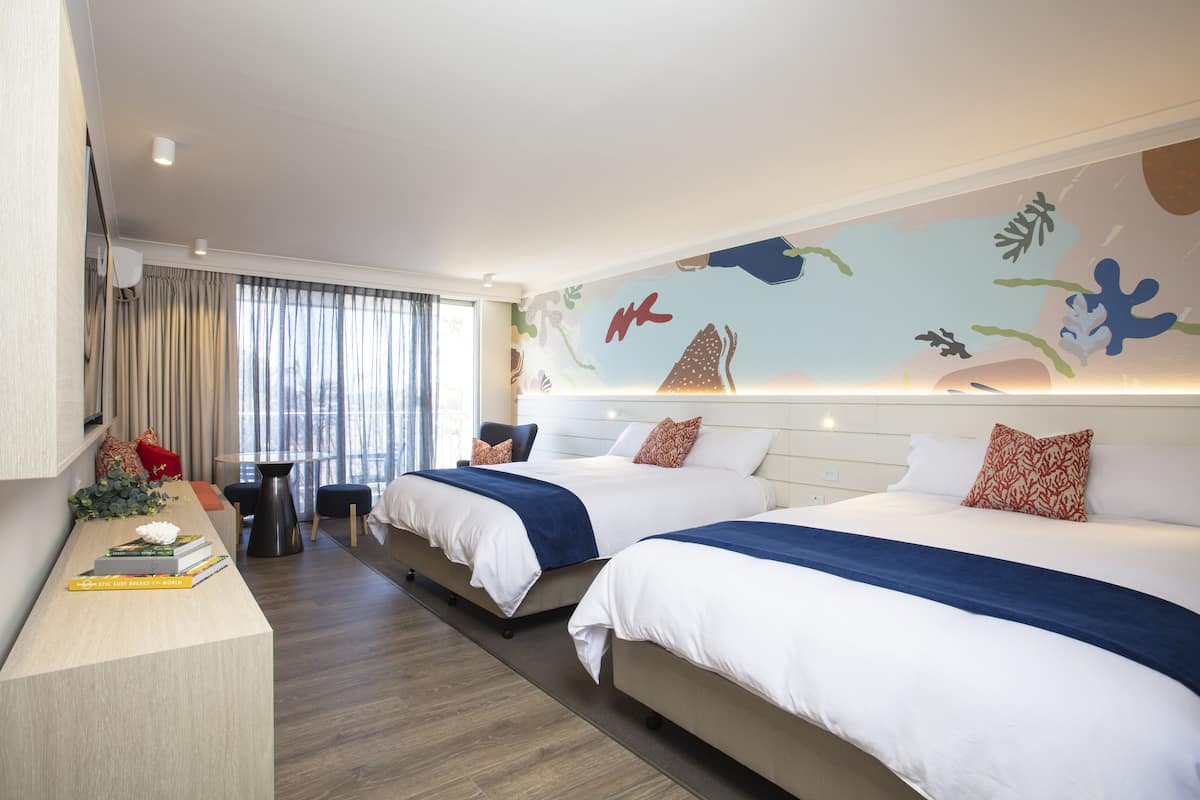 A hotel room with two beds, a desk with books, a small table, and a mural on the wall.