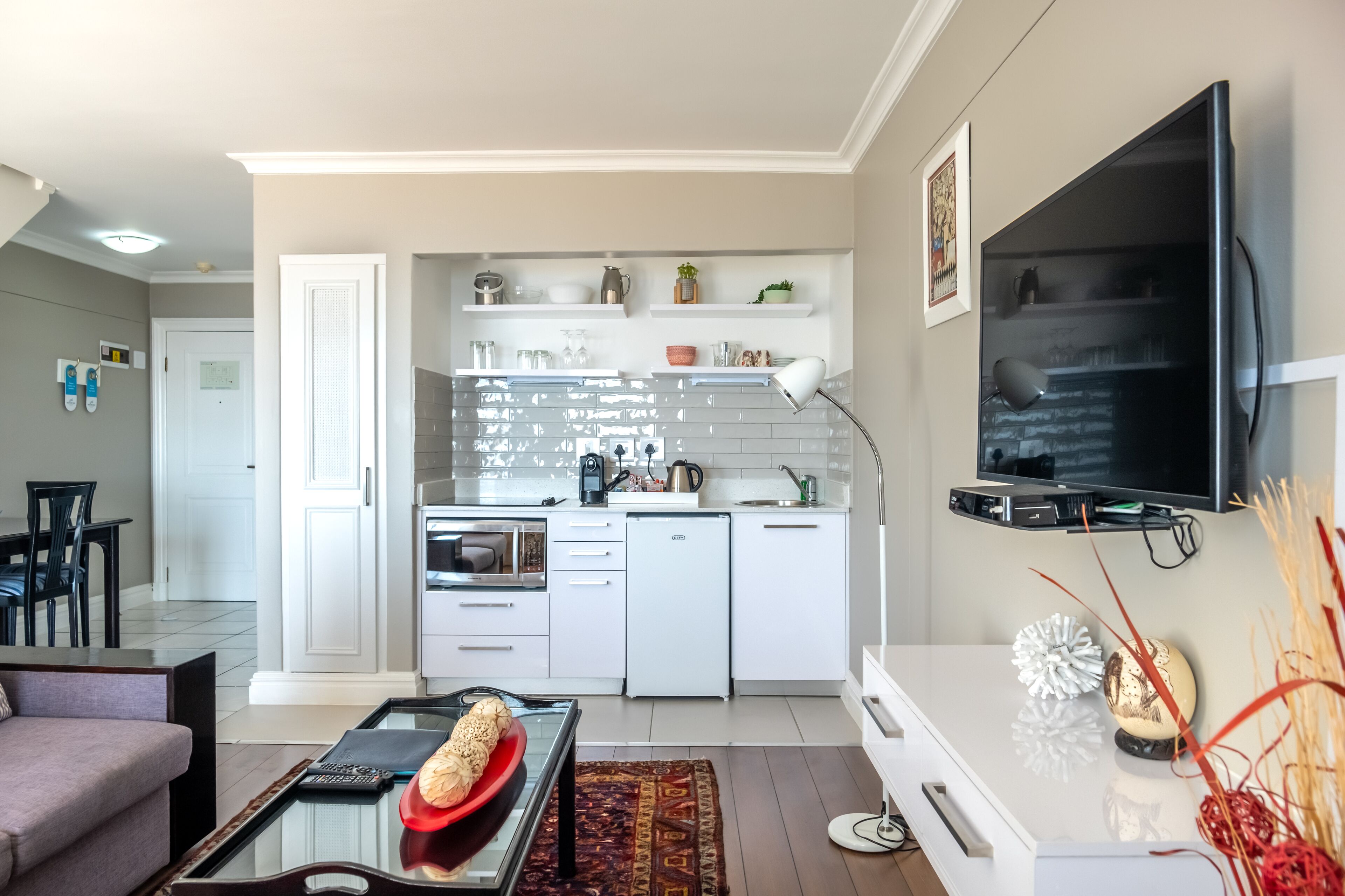 A modern kitchen with white cabinets, a built-in oven, and a microwave. There is a dining area with a table and chairs, and a living room with a sofa and a coffee table.