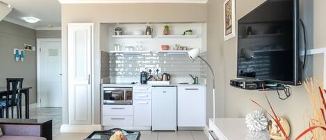 A modern kitchen with white cabinets, a built-in oven, and a microwave. There is a dining area with a table and chairs, and a living room with a sofa and a coffee table.