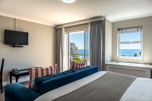 A hotel room with a bed, a flat-screen TV, a desk with a telephone, and a view of the sea through large windows.