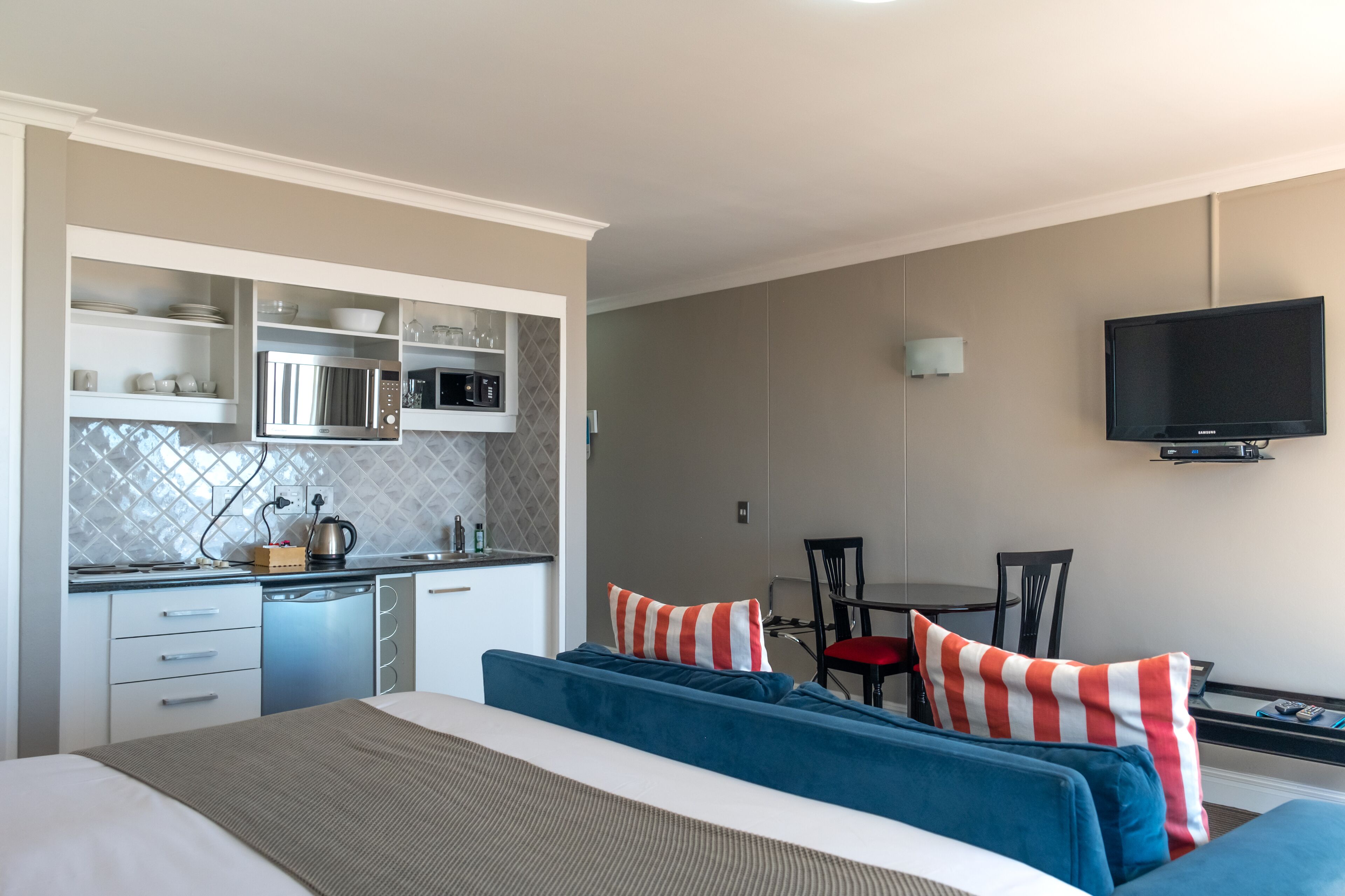 Ein kompaktes Hotelzimmer mit Küchenzeile, einem Bett mit blauer Bettwäsche, einem an der Wand befestigten Fernseher und einem Essbereich mit Tisch und Stühlen.