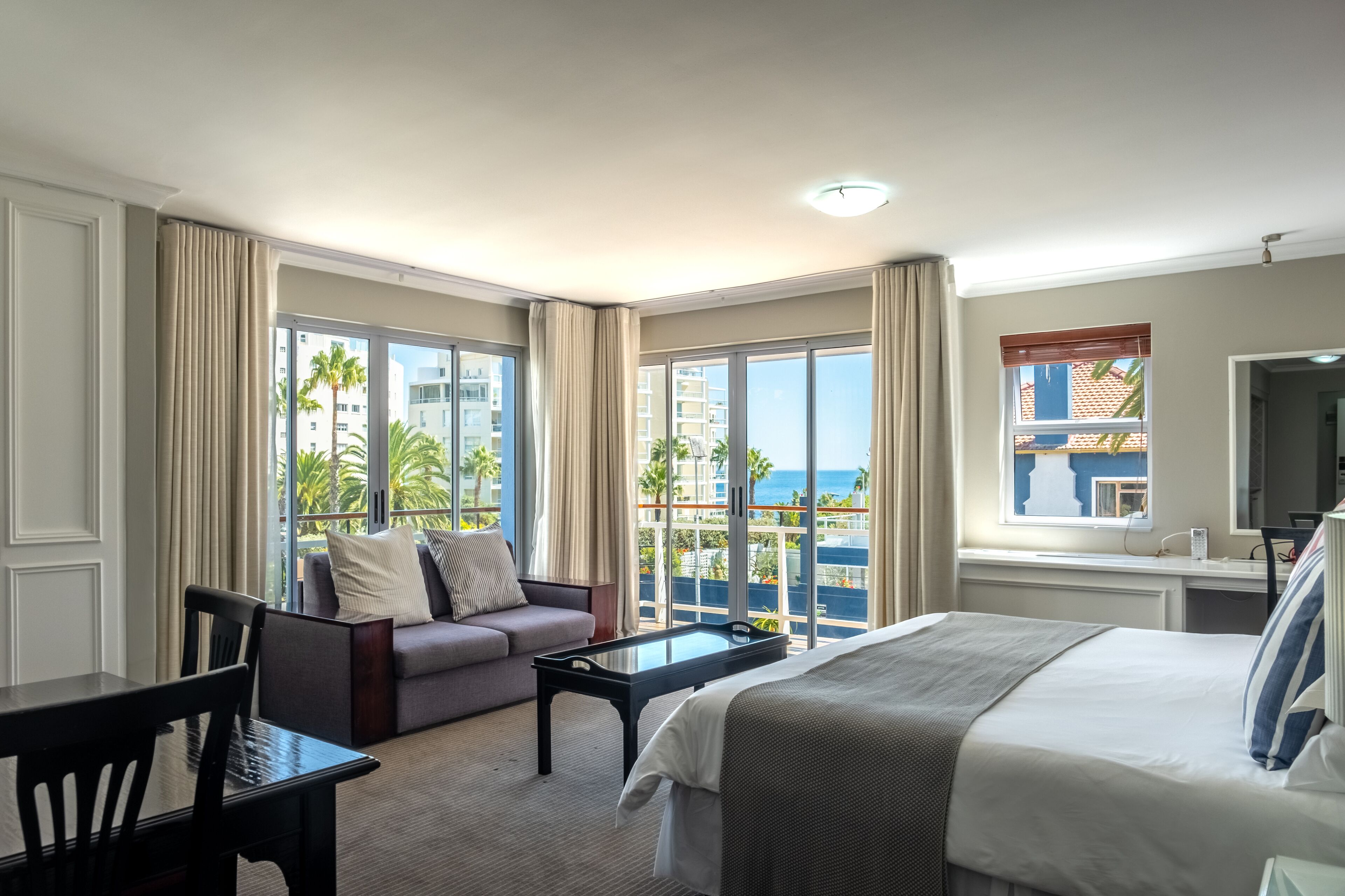 Habitación de hotel con cama, sofá, mesa de comedor y un amplio ventanal con vista al océano y edificios.