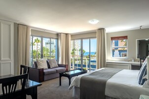 A hotel room with a bed, a sofa, a dining table, and a large window offering a view of the ocean and buildings.