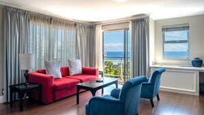 A modern living room with a red sofa, blue armchair, and a view of the ocean through large windows. - The Bantry Bay Aparthotel by Totalstay (Cape Town)