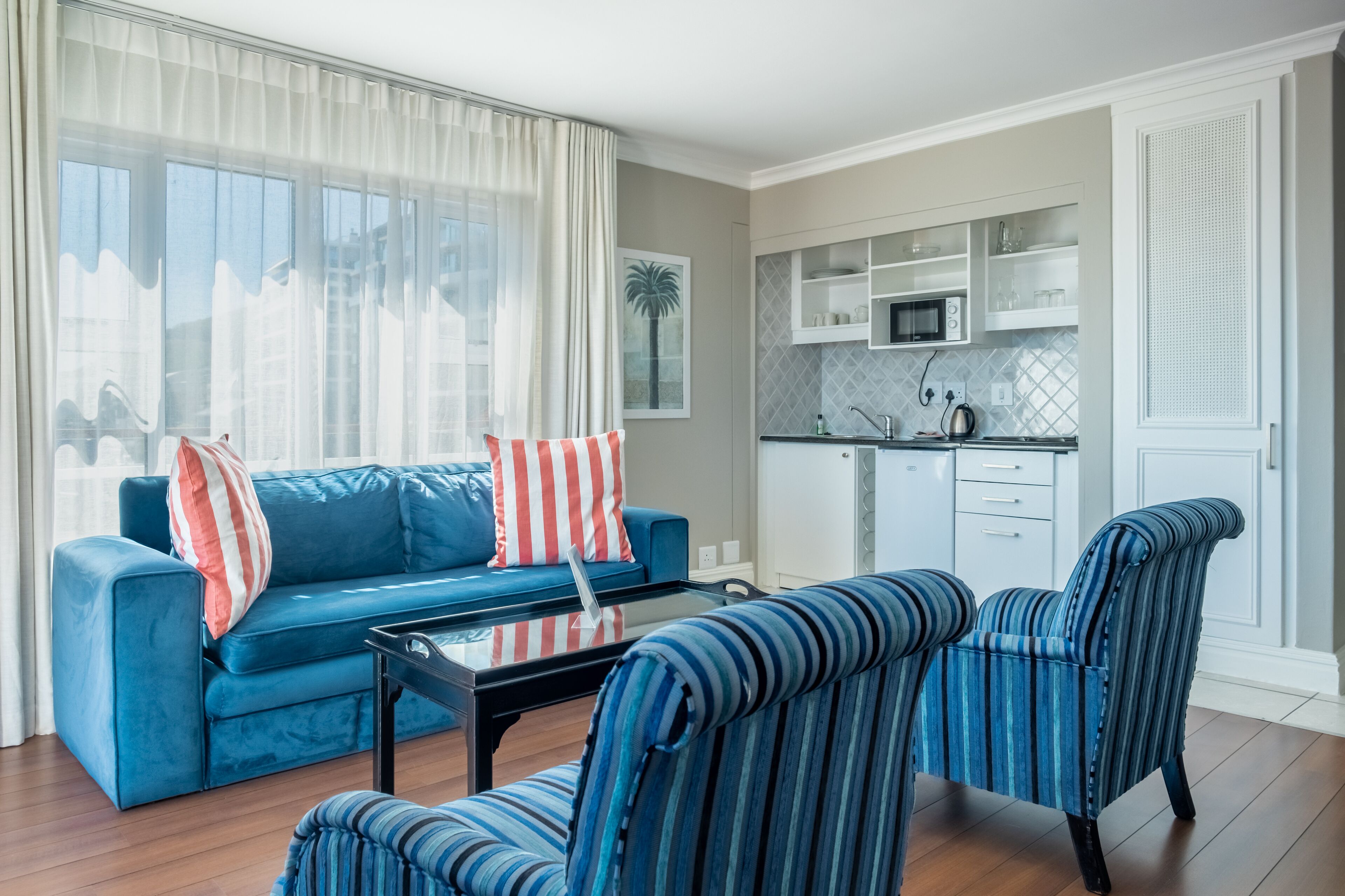 Een moderne woonkamer met een blauwe bank, gestreepte fauteuils, een glazen salontafel en een keuken met witte kasten en een magnetron.
