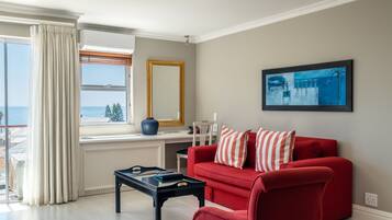 A living room with a red sofa, a black coffee table, a large window with curtains, a painting on the wall, and a view of the sea.