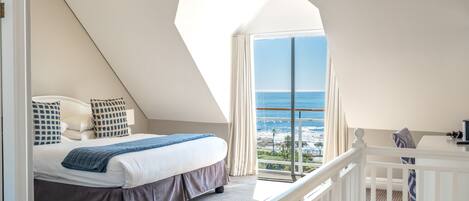 Een slaapkamer met een groot raam dat uitkijkt over de zee, een bed met een blauwe deken en een witte balustrade.
