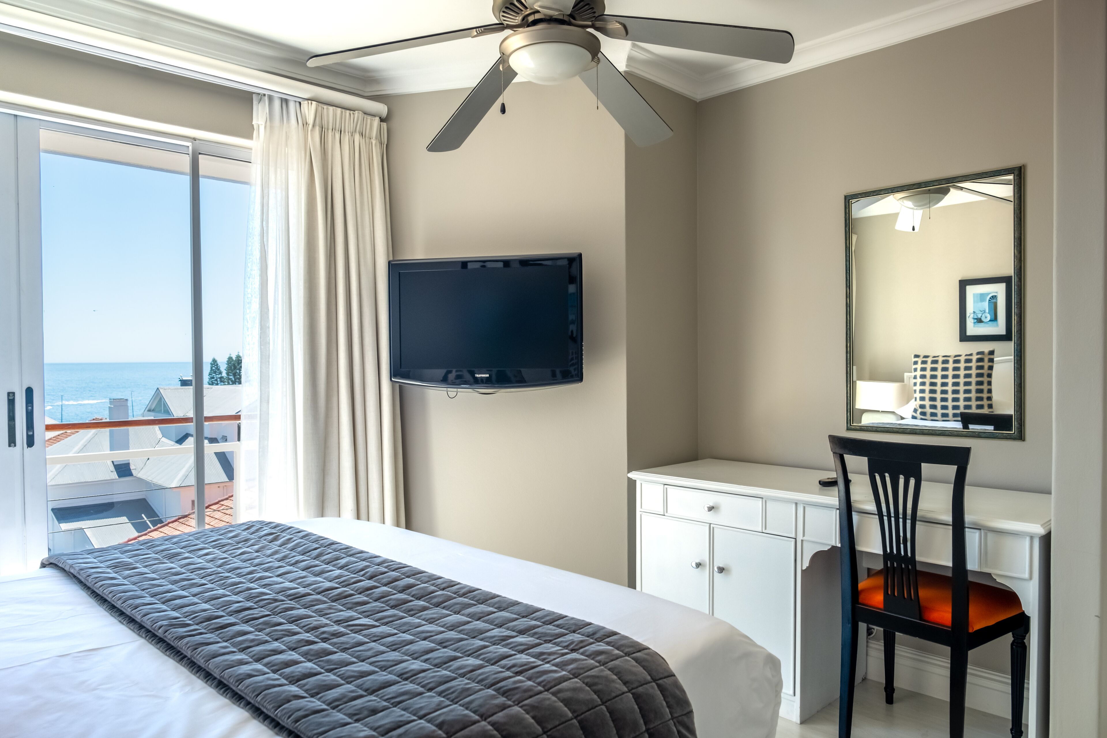 A hotel room with a bed, a desk, a chair, a television, and a balcony with a view of the sea.