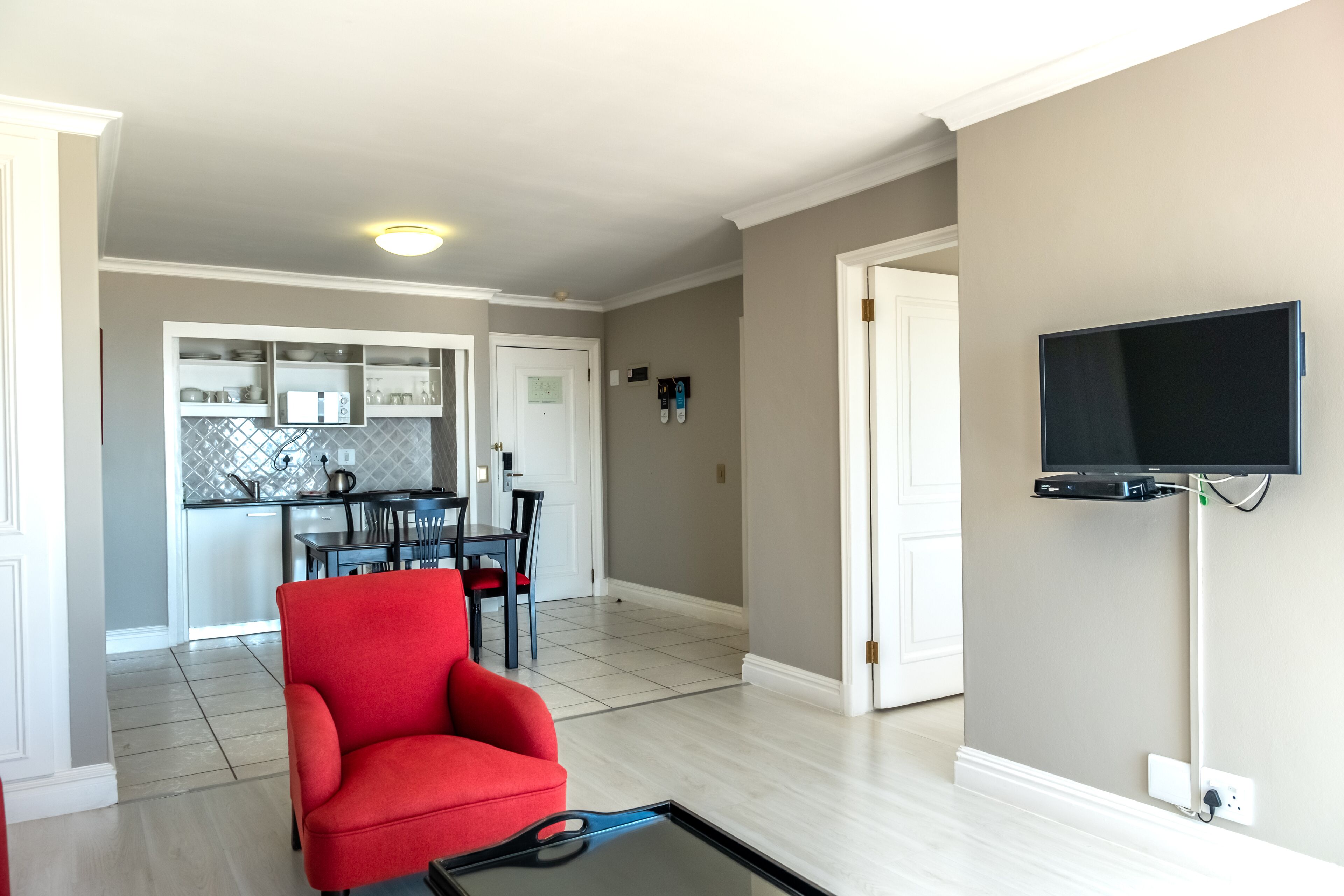 Un salon avec un fauteuil rouge, un téléviseur à écran plat fixé au mur et un coin cuisine à l’arrière-plan.