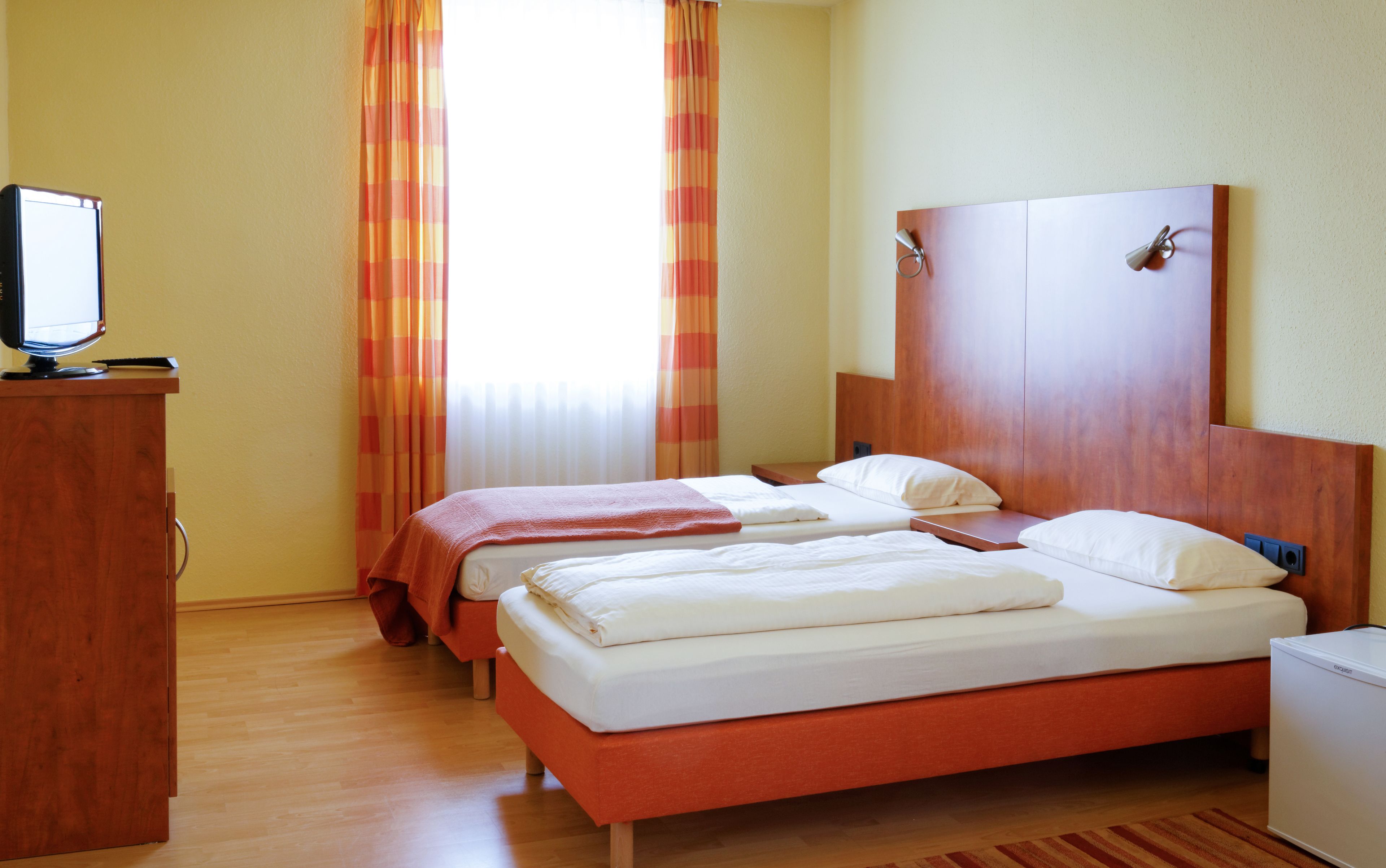 A hotel room with two beds, a wooden headboard, a small desk with a computer, and a window with curtains.