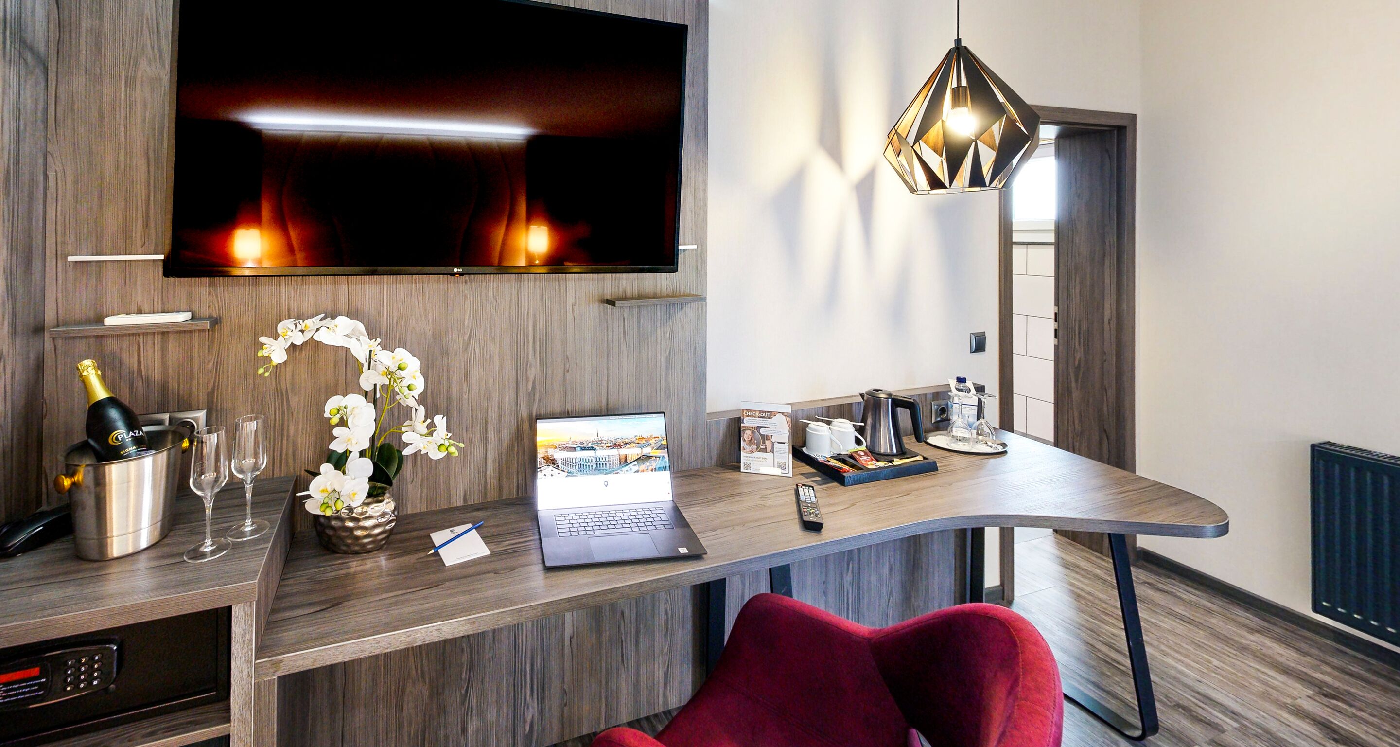 A modern hotel room with a flat-screen TV, a laptop, a minibar, a red chair, and a wooden desk.