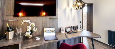 A modern hotel room with a flat-screen TV, a laptop, a minibar, a red chair, and a wooden desk.