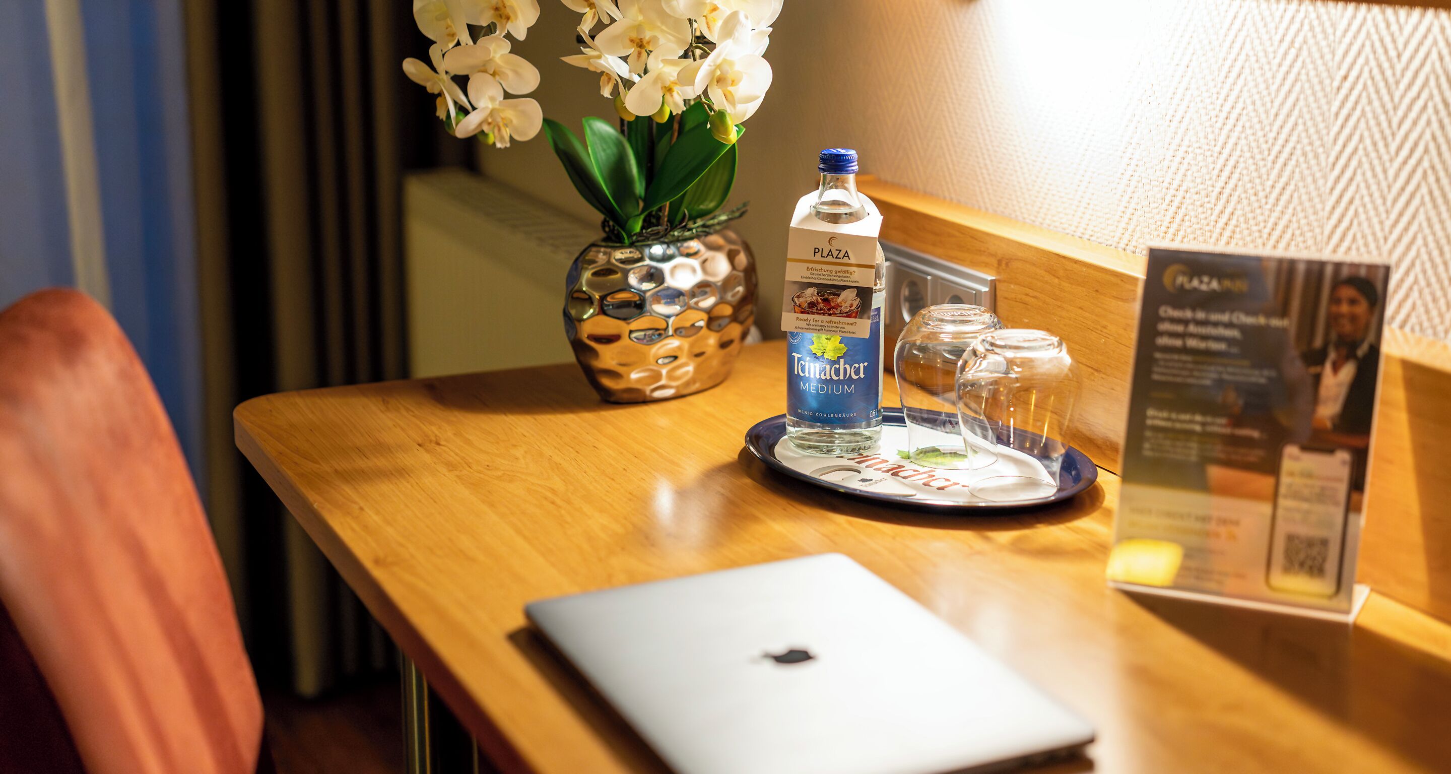 A wooden table with a laptop, a bottle of water, a glass, and a vase with flowers.