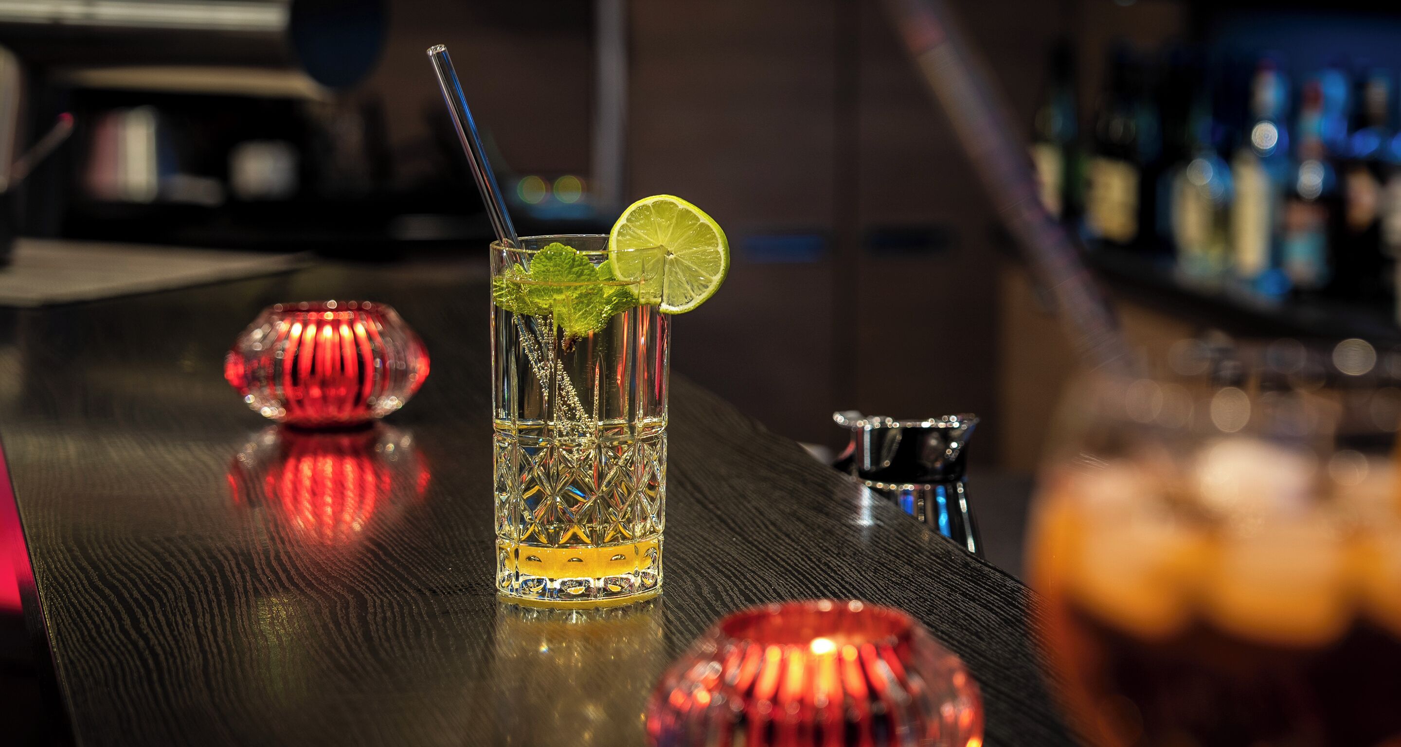 Un bar avec un verre contenant une boisson décorée d’une tranche de citron vert et de feuilles de menthe, un objet décoratif rouge et un récipient en métal posé sur le comptoir.