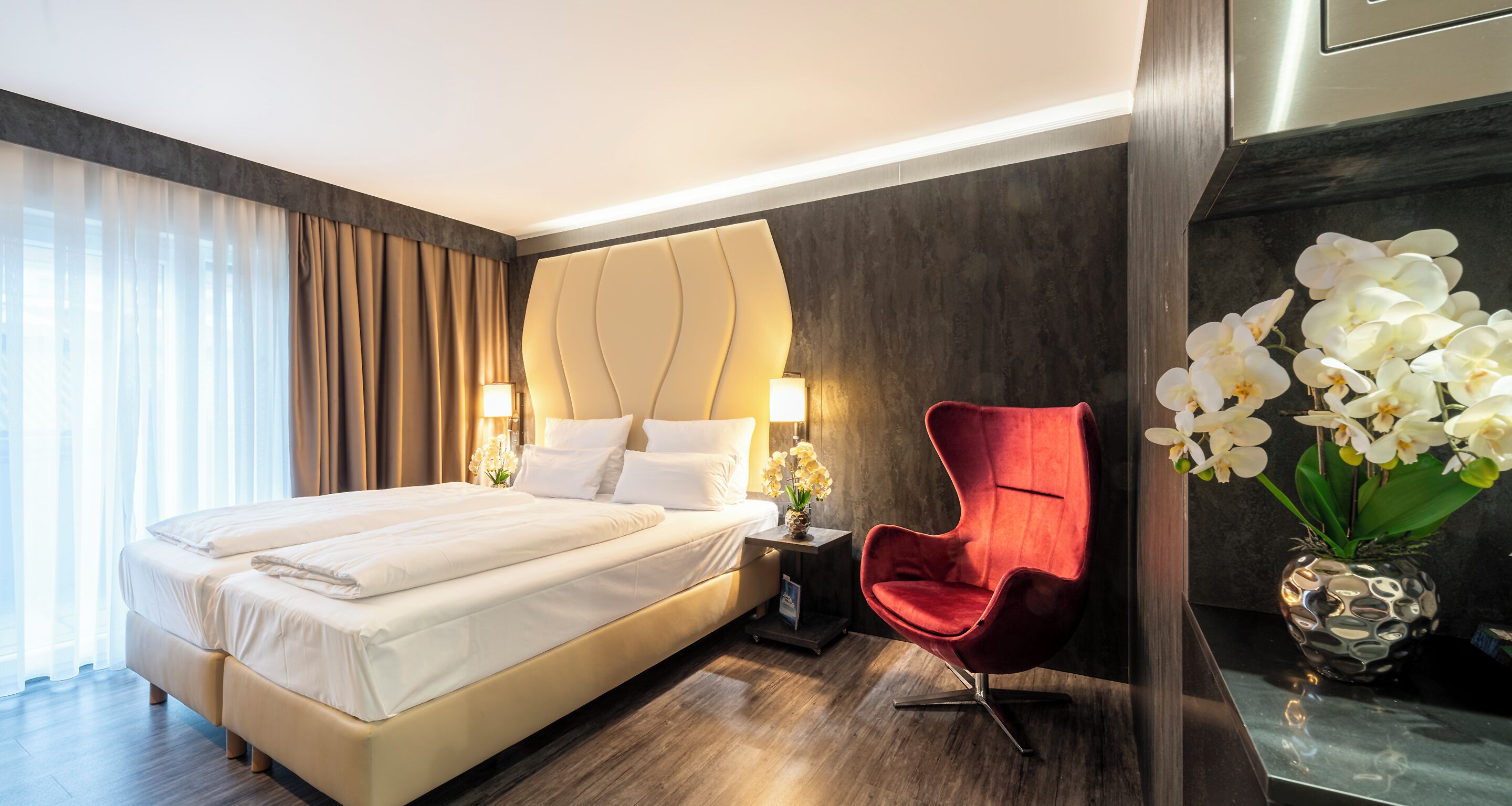 A hotel room with a large bed, a red chair, a vase with white flowers, and a modern wall design.
