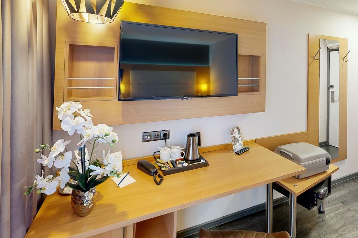 A hotel room with a flat-screen TV mounted on the wall, a wooden desk with a vase of white flowers, a coffee maker, a telephone, and a small seating area.
