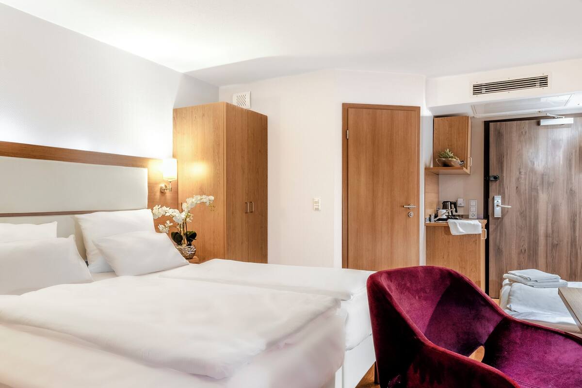 A hotel room with a large bed, a wooden wardrobe, a vanity with a mirror, and a red chair.