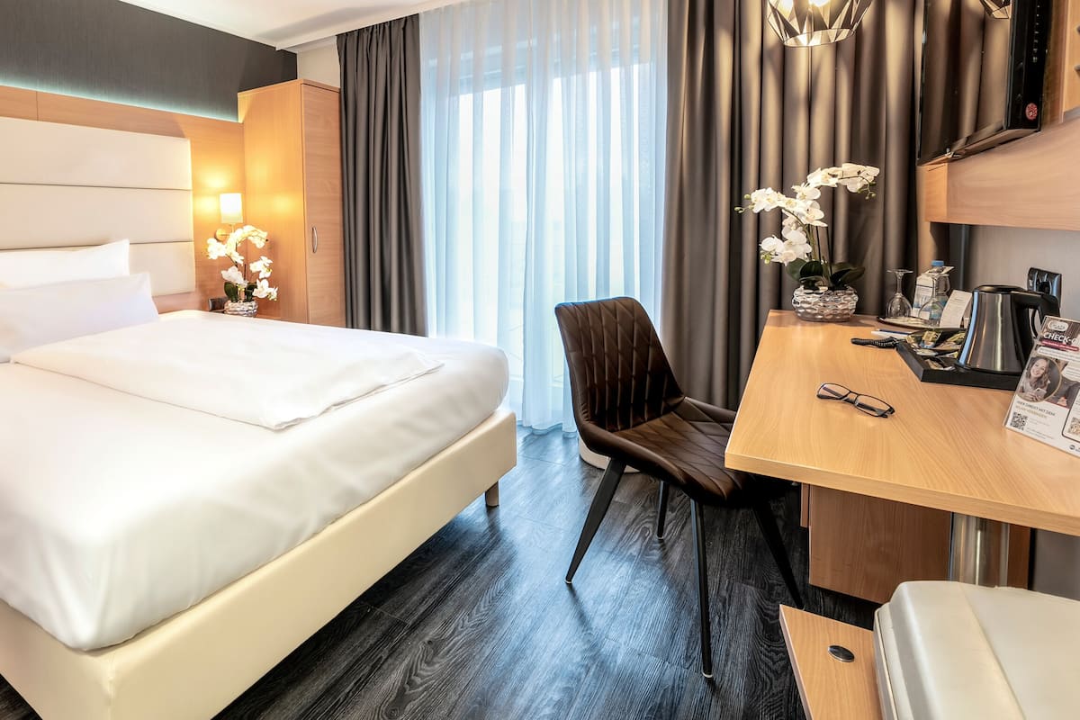 A hotel room with a bed, a desk, a chair, a nightstand, and a lamp.