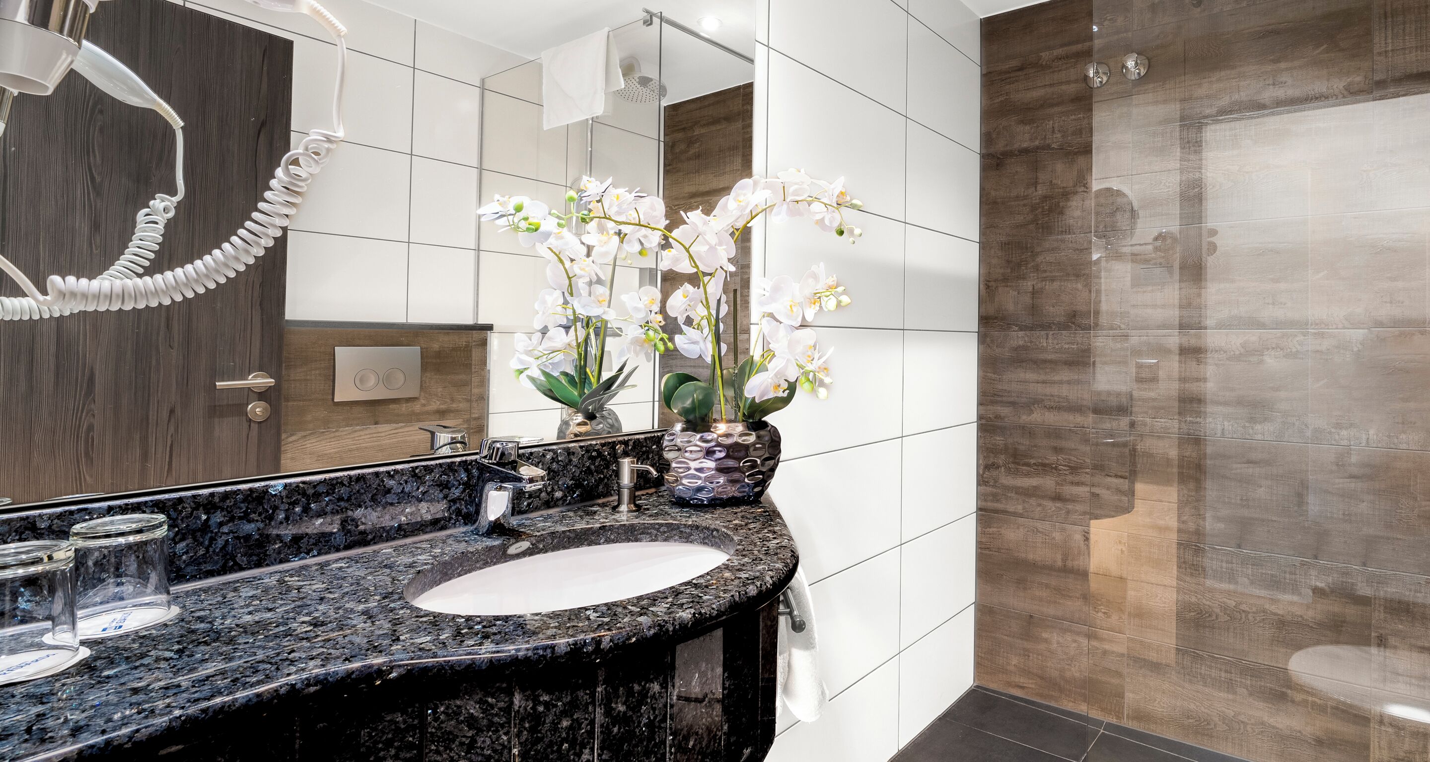 A modern bathroom with a granite countertop, a white sink, a large mirror, and a shower with a wooden panel design.