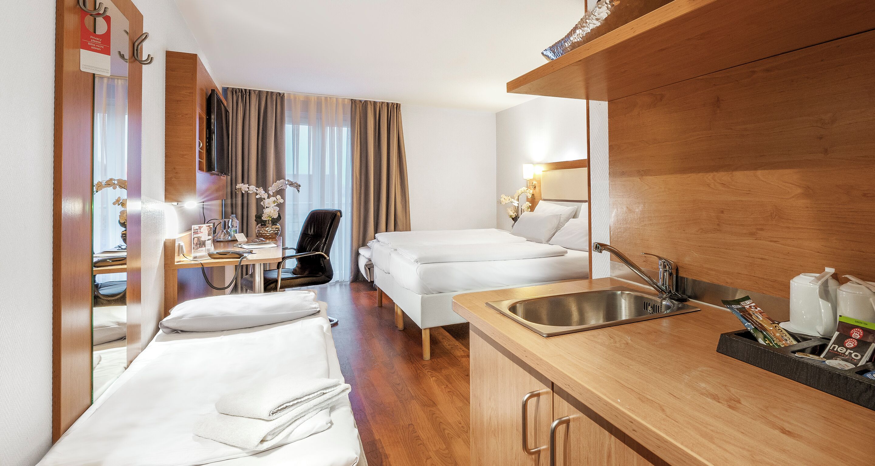 Une chambre d’hôtel comprenant un lit, un bureau avec une chaise, une petite kitchenette avec un évier et un miroir.