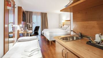 Habitación de hotel con cama, escritorio con silla, kitchenette pequeña con lavabo y espejo.