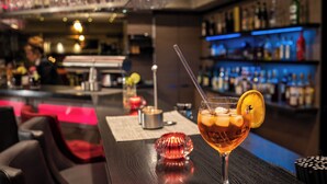 A bar area with a cocktail glass on a wooden bar counter, a red chair, and a bar stocked with bottles. - PLAZA INN Heilbronn (Heilbronn)
