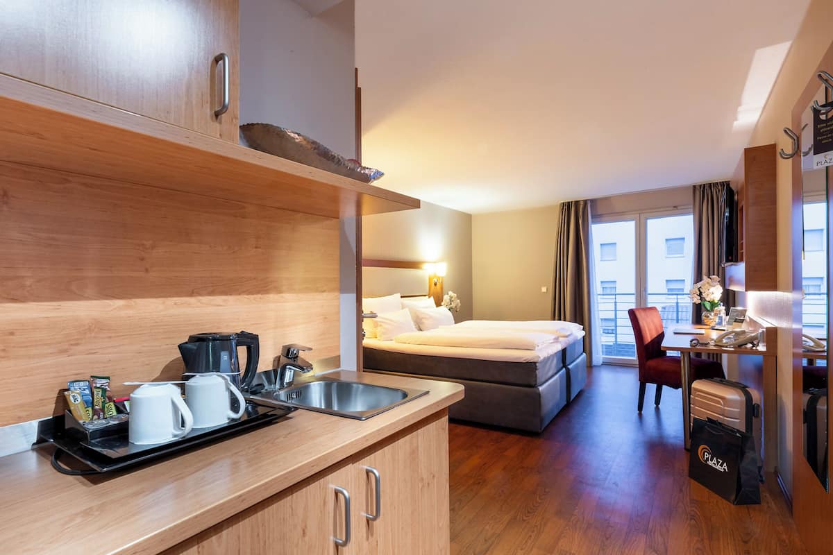A compact hotel room with a kitchenette, a bed, a desk, and a view of the city.
