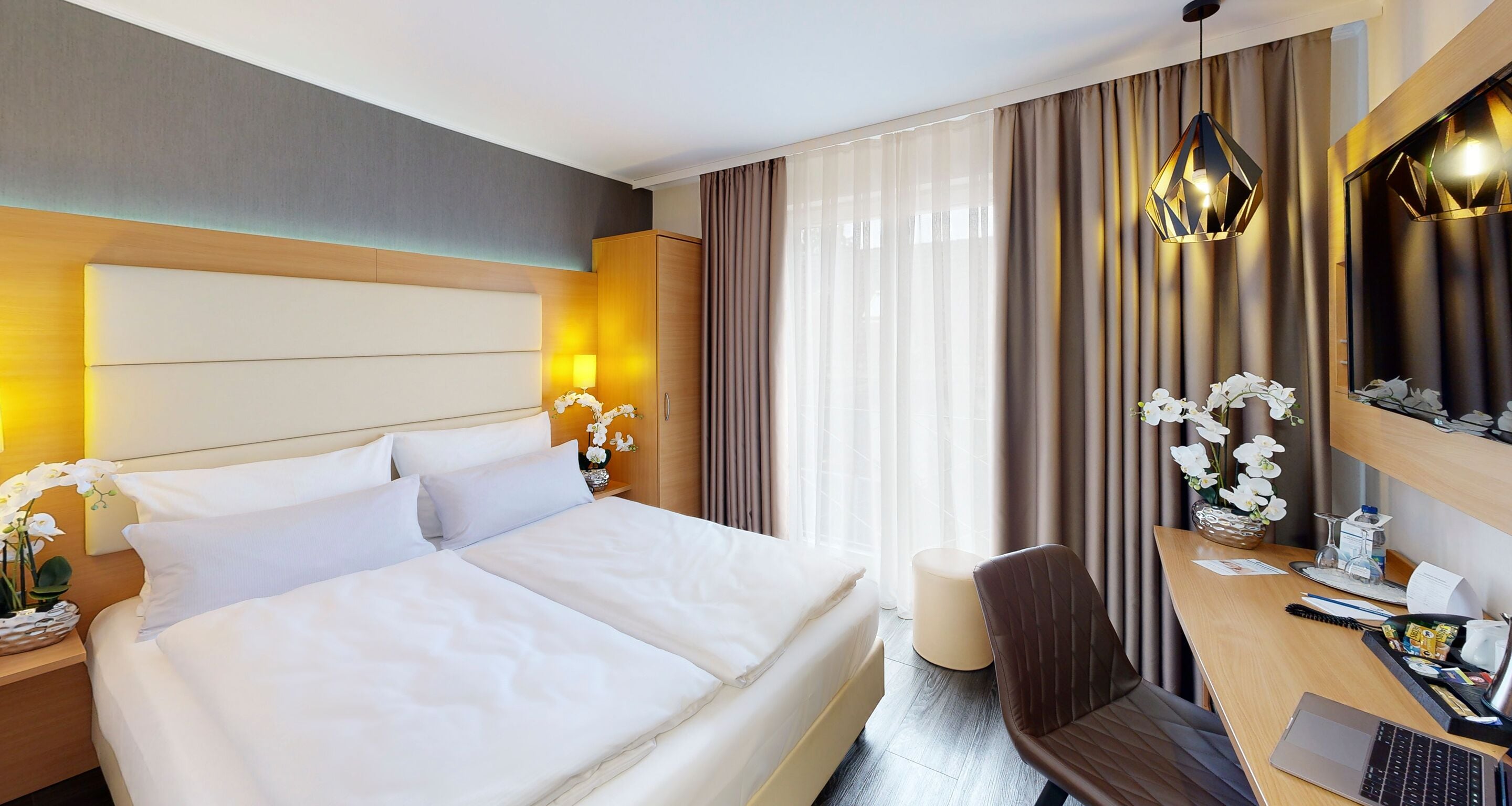 A hotel room with a large bed, a desk with a laptop, a chair, and a small table with a vase of flowers.