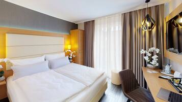 A hotel room with a large bed, a desk with a laptop, a chair, and a small table with a vase of flowers.