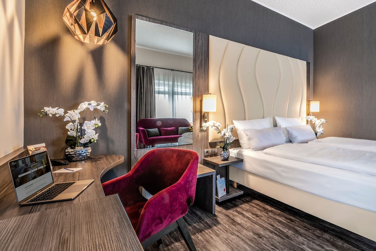 A hotel room with a bed, a desk with a laptop, a red chair, a mirror, and a vase with flowers.