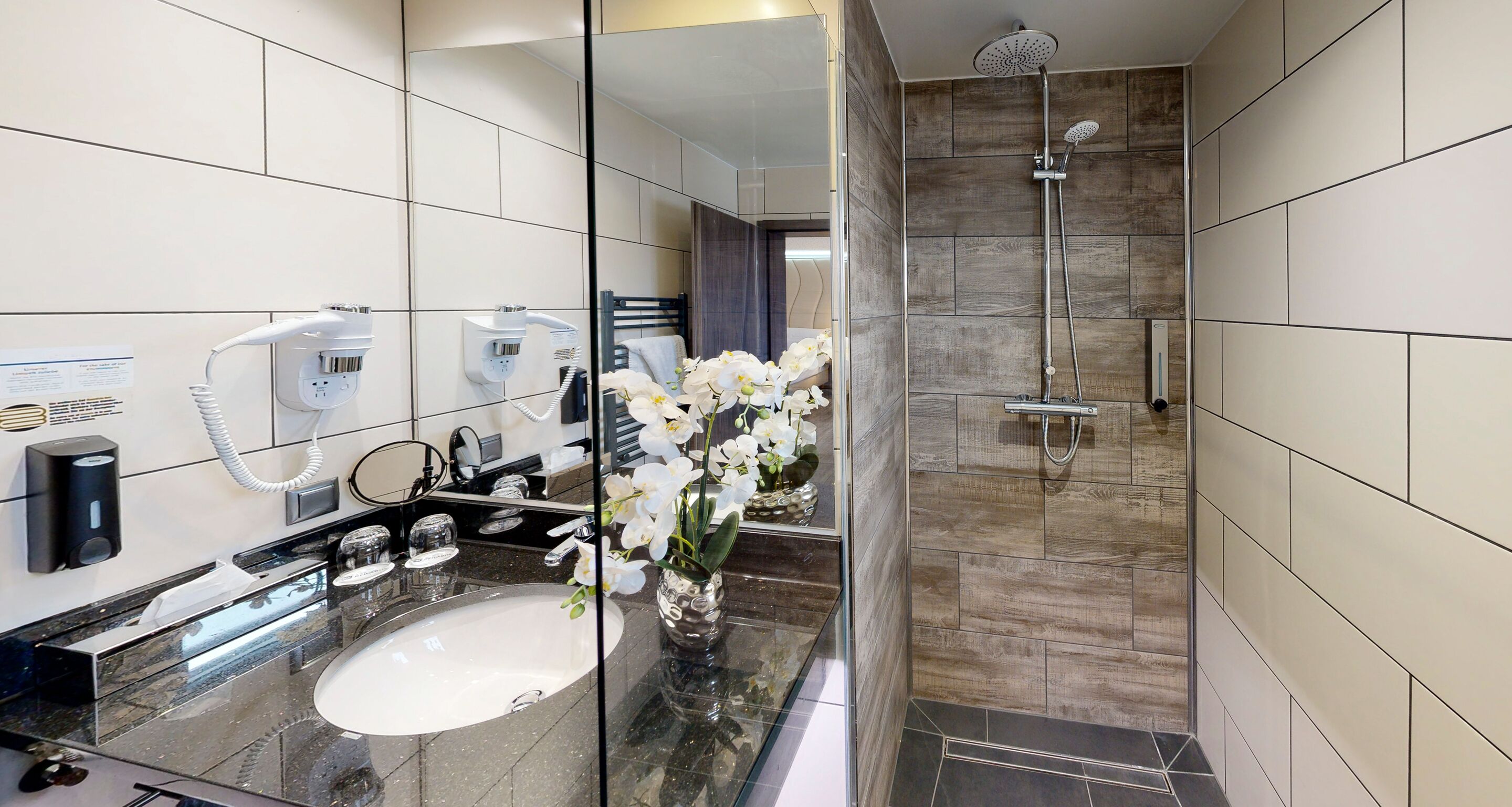 Un baño moderno con un espejo grande, un lavamanos, una ducha y un florero con flores blancas.