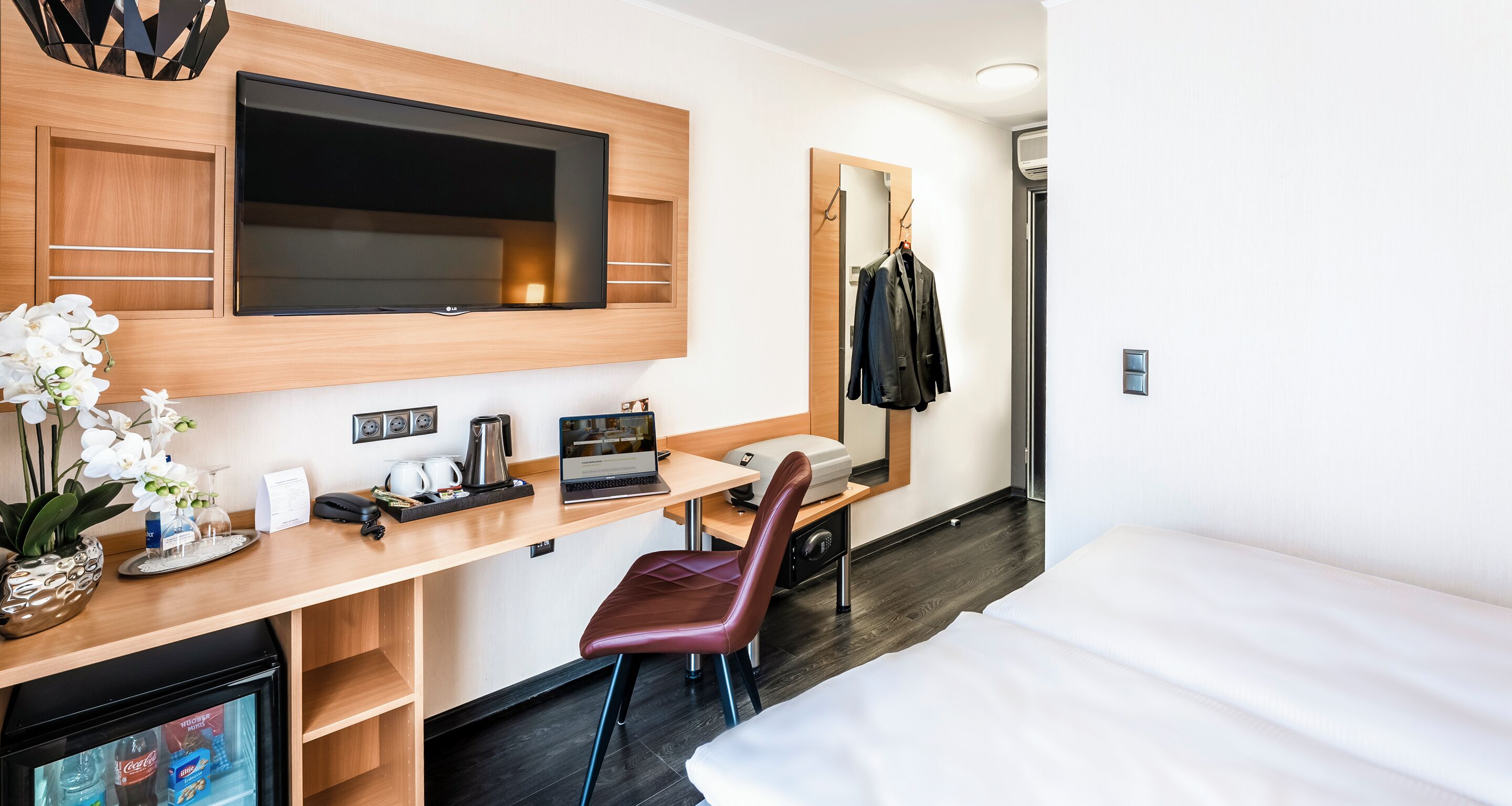 Ein Hotelzimmer mit einem Bett, einem Schreibtisch mit einem Laptop, einem Stuhl, einem an der Wand befestigten Fernseher, einem Spiegel und einem Kleiderhaken.