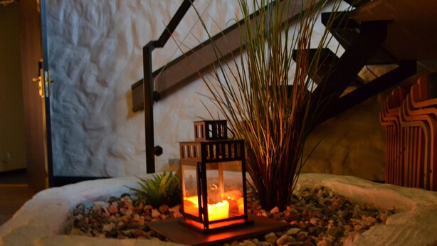 Una jardinera de piedra con un farol iluminado, rodeada de piedras y plantas, frente a una pared con textura.
