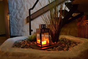 Una jardinera de piedra con un farol iluminado, rodeada de piedras y plantas, frente a una pared con textura.