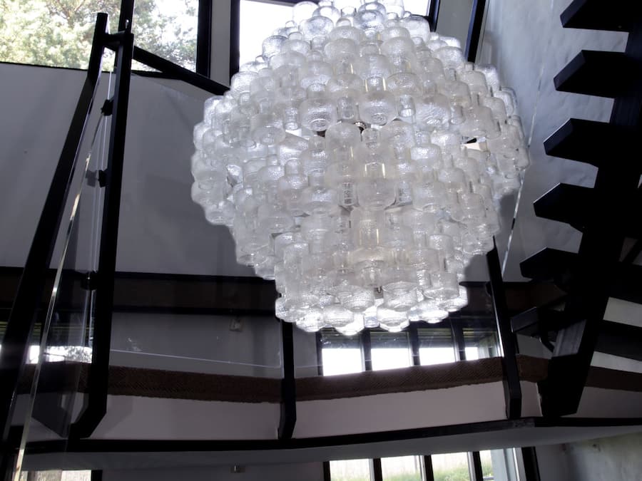 Un candelabro hecho de botellas de vidrio transparente que cuelga en un vestíbulo con ventanas amplias.