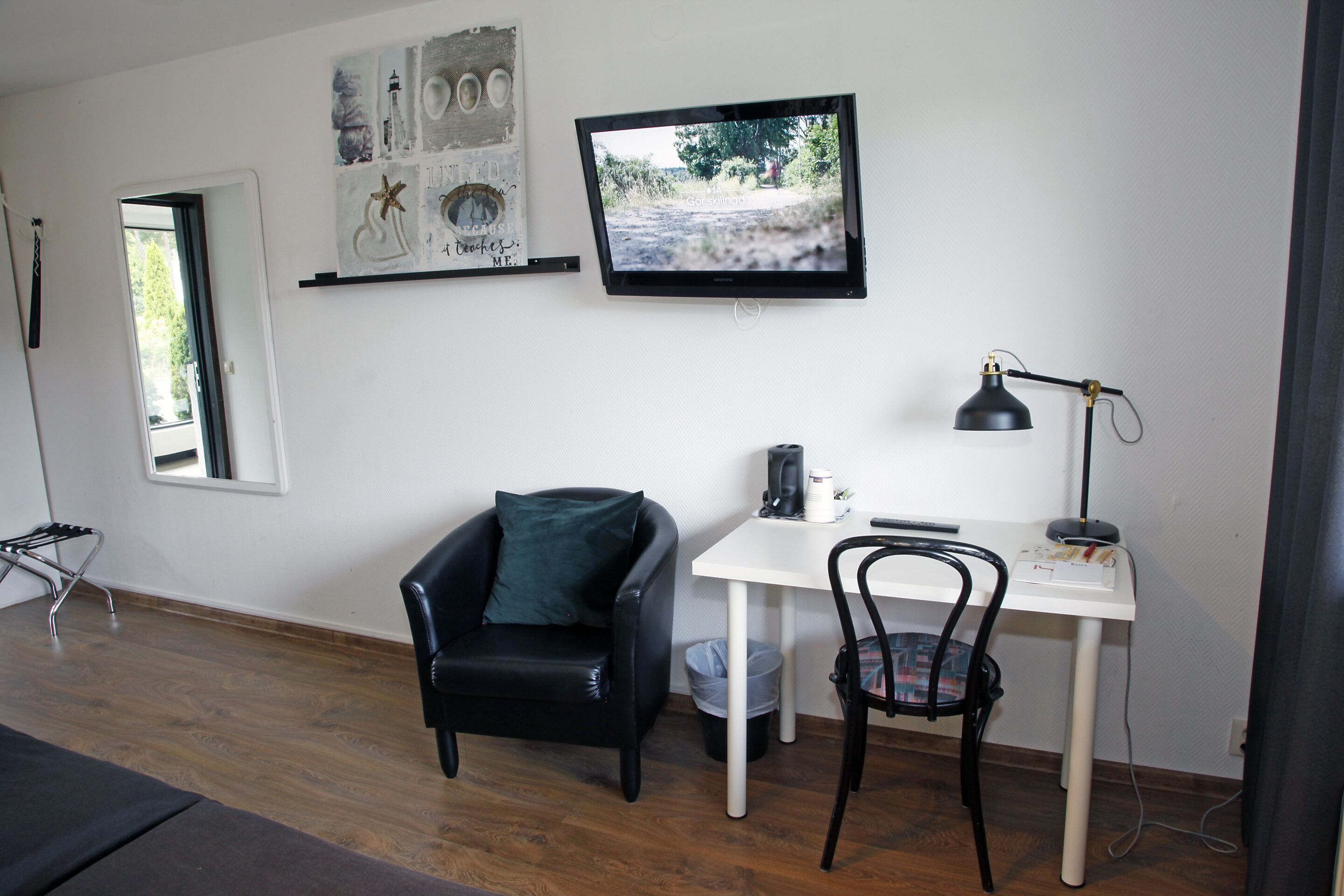 A room with a white desk, a black chair, a black leather sofa, a television mounted on the wall, and a small window.