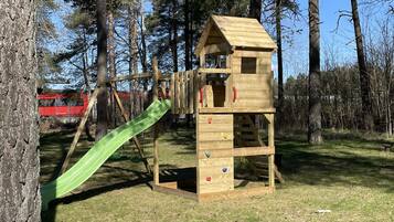A wooden play structure with a slide and climbing wall in a grassy area with trees.