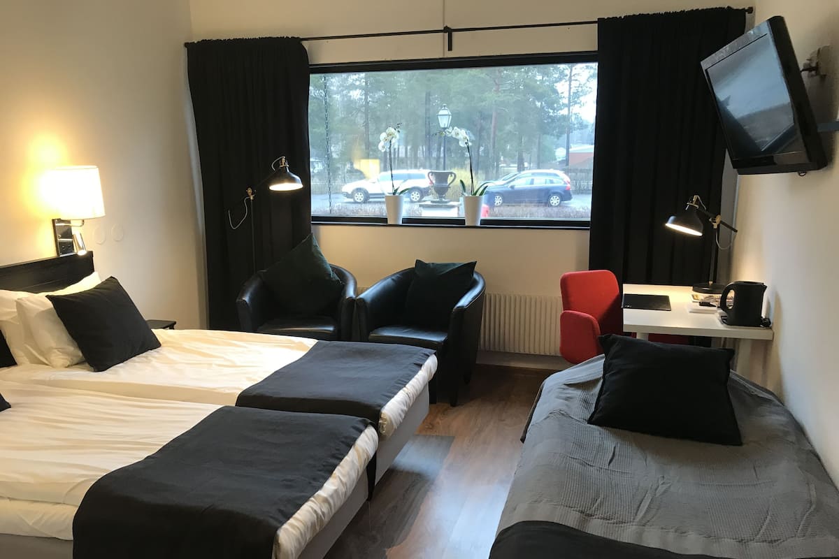 A hotel room with two beds, a desk, a chair, a TV, and a window with a view of cars and trees.