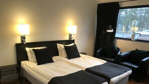 Habitación de hotel con cama doble, dos mesitas de noche, una silla y ventana con vistas a árboles y un coche.