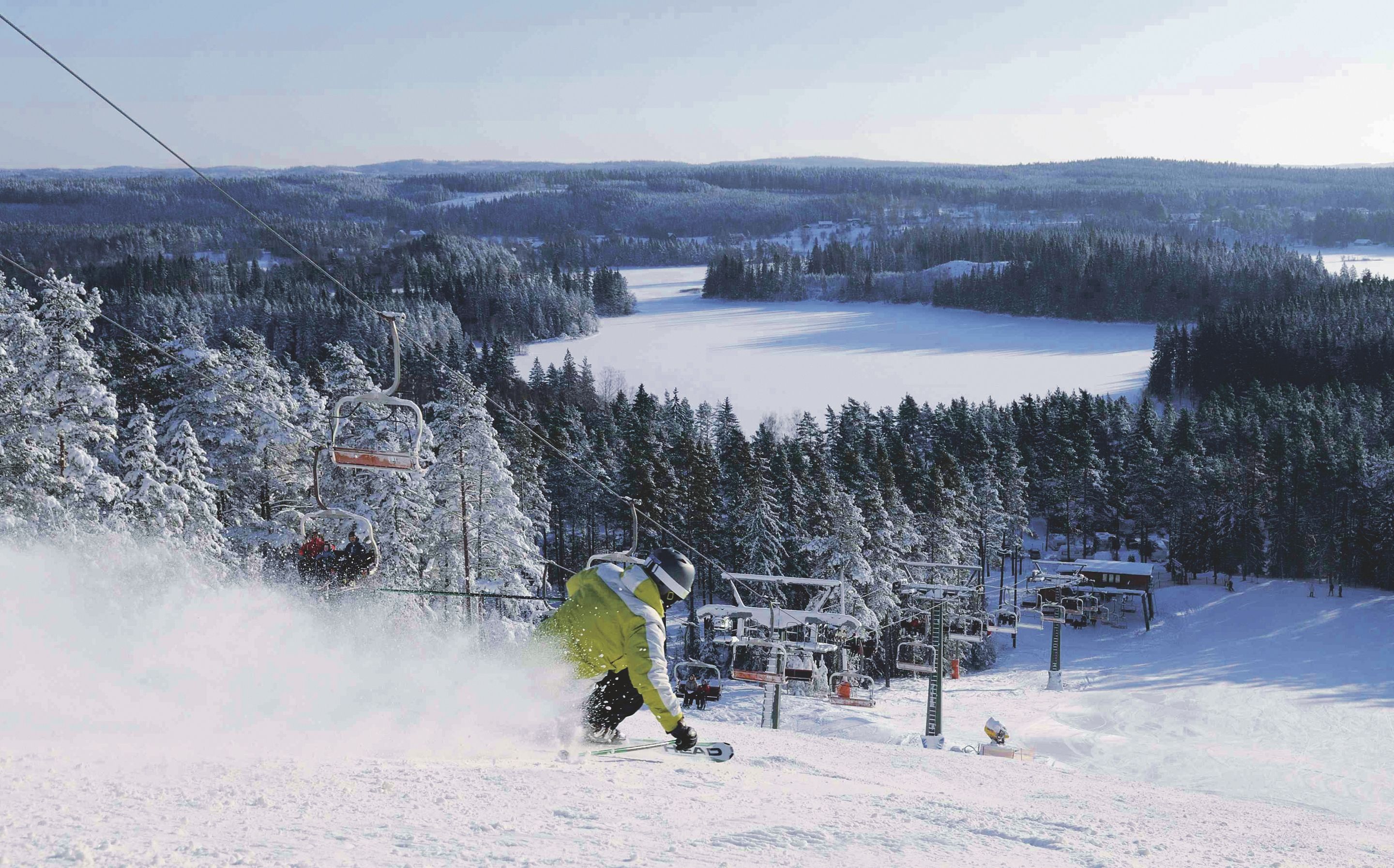 Eine Skipiste mit einer Person, die abfährt, umgeben von schneebedeckten Bäumen und einem See im Hintergrund.