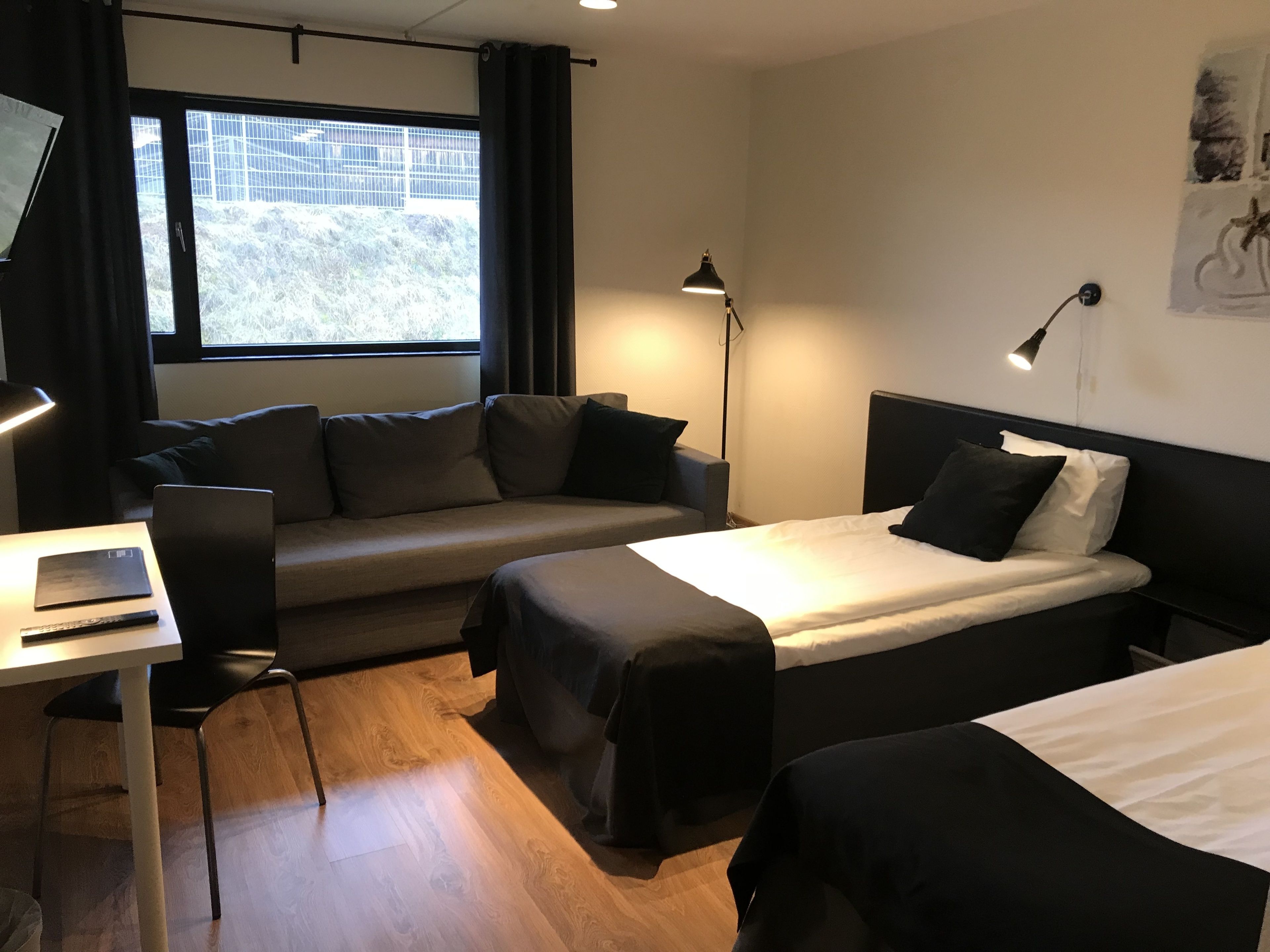 Ett hotellrum med en säng, ett skrivbord med en bärbar dator, en stol, en soffa och ett fönster med gardiner.