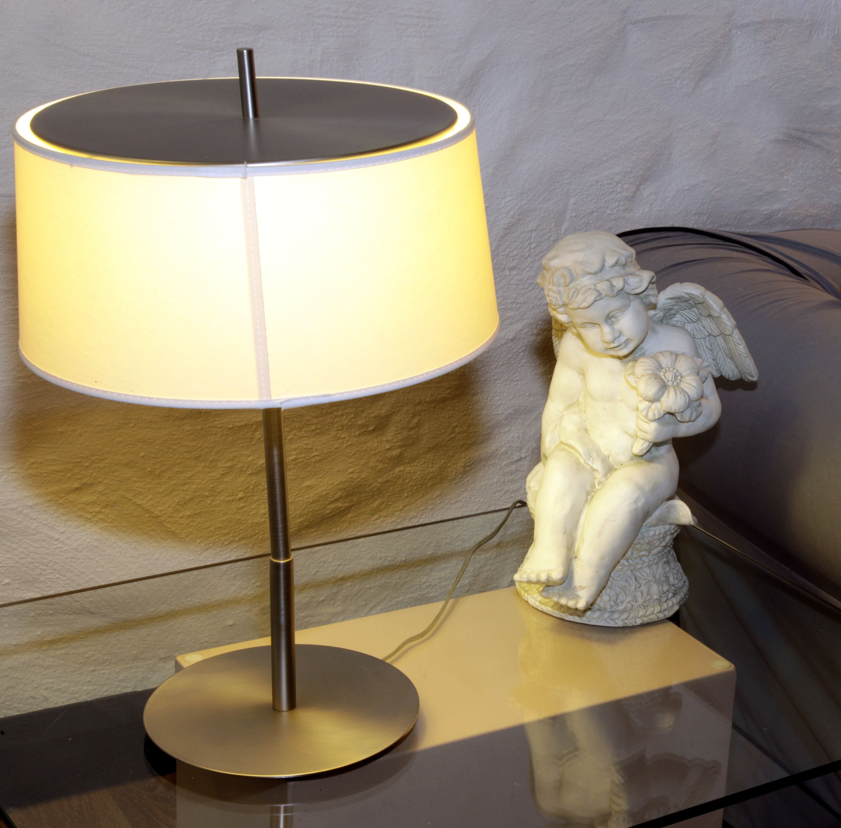 Een bureaulamp met een witte kap en een metalen voet, geplaatst naast een wit beeldje van een cherubijn op een glazen tafel.
