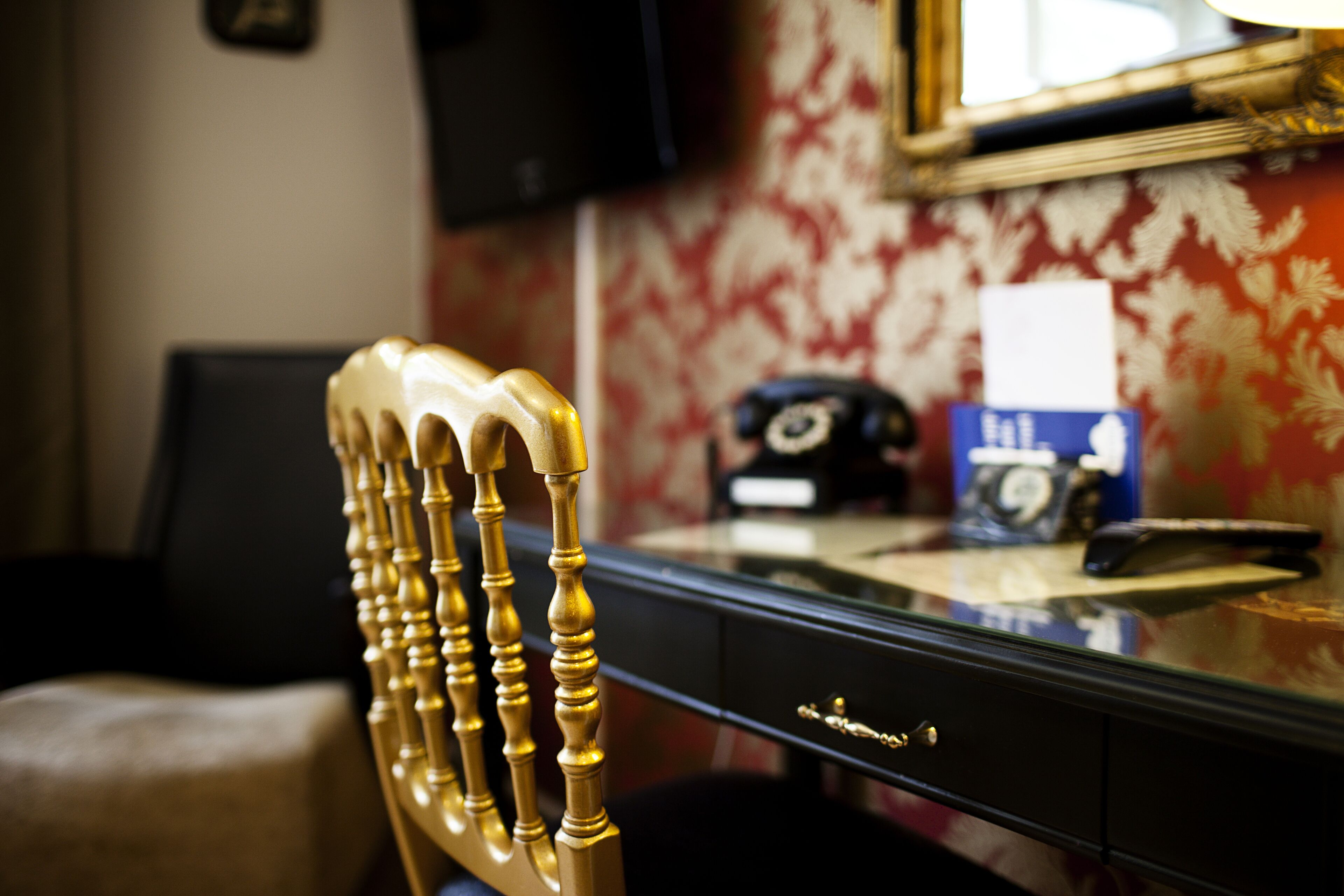 A room with a wooden chair with a curved backrest, a black desk with a telephone and a blue box, and a mirror on the wall.
